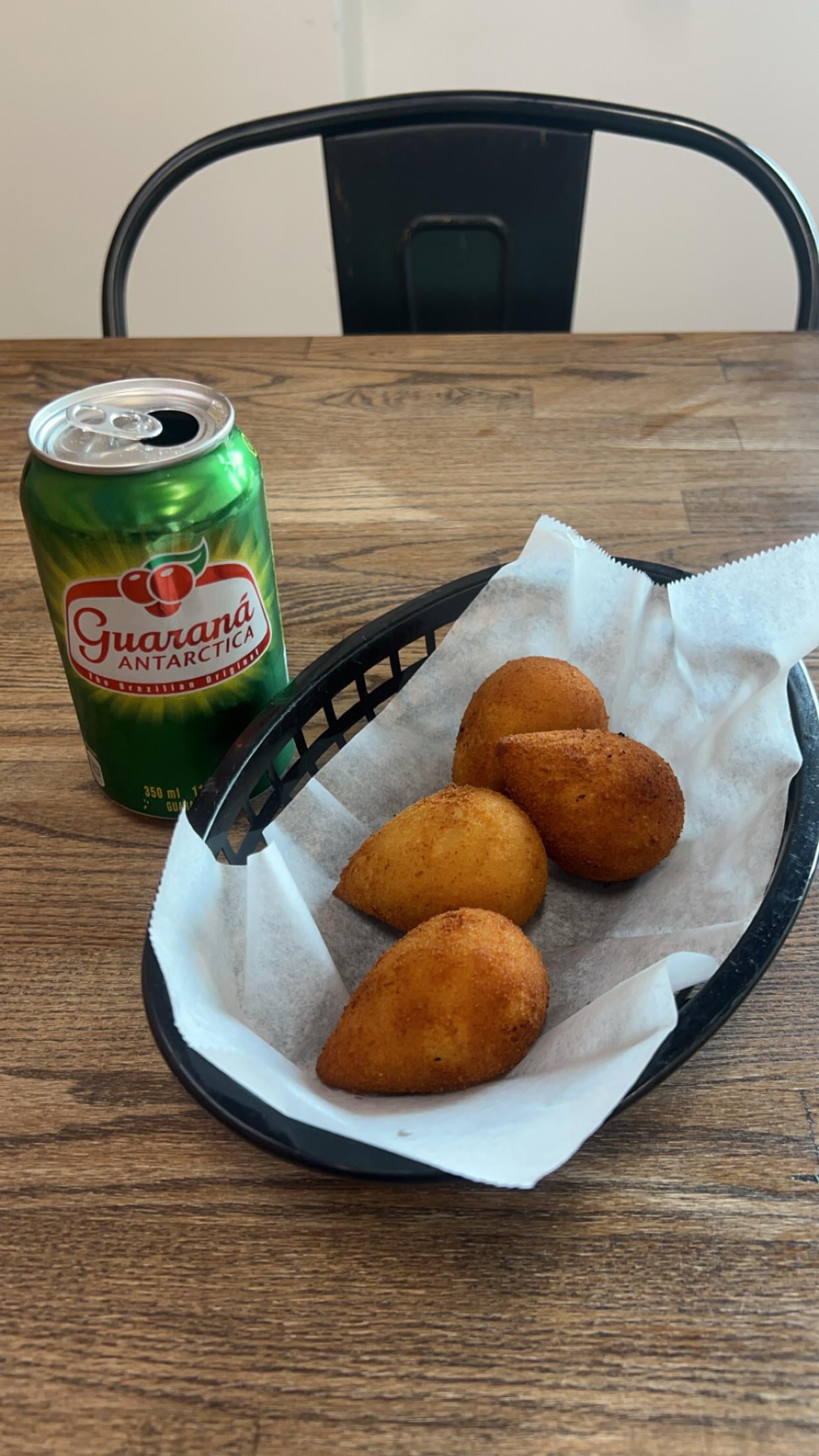 A green can of Guaraná Antarctica soda sits next to a black basket lined with white paper, containing four golden-brown coxinhas on a wooden table. A black metal chair is in the background.