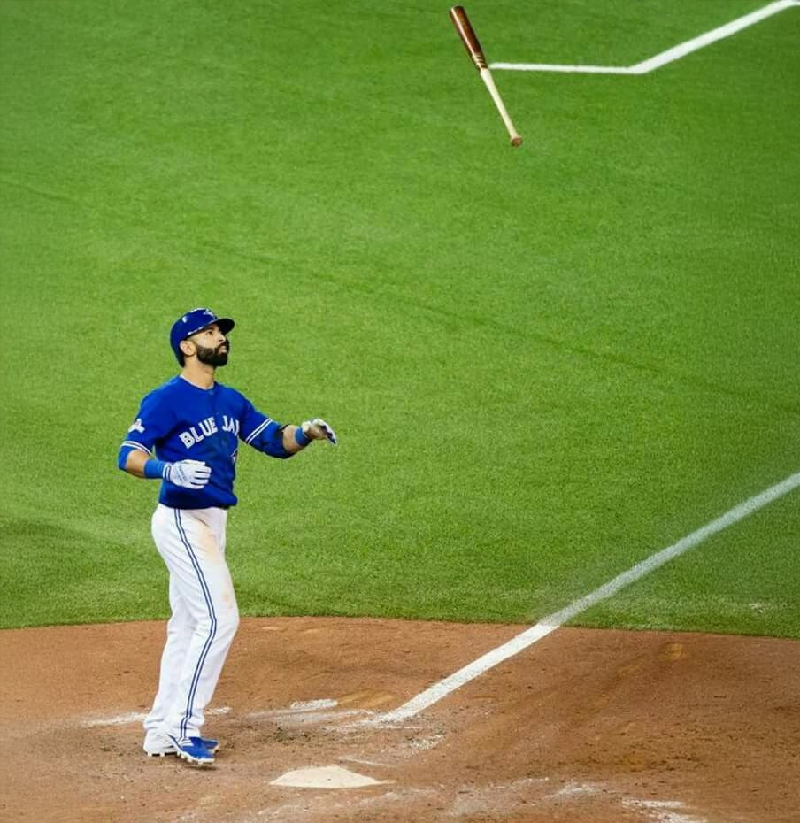 A baseball player in a Blue Jays uniform stands at home plate, looking up as a wooden bat spins in the air above him on a green baseball field.