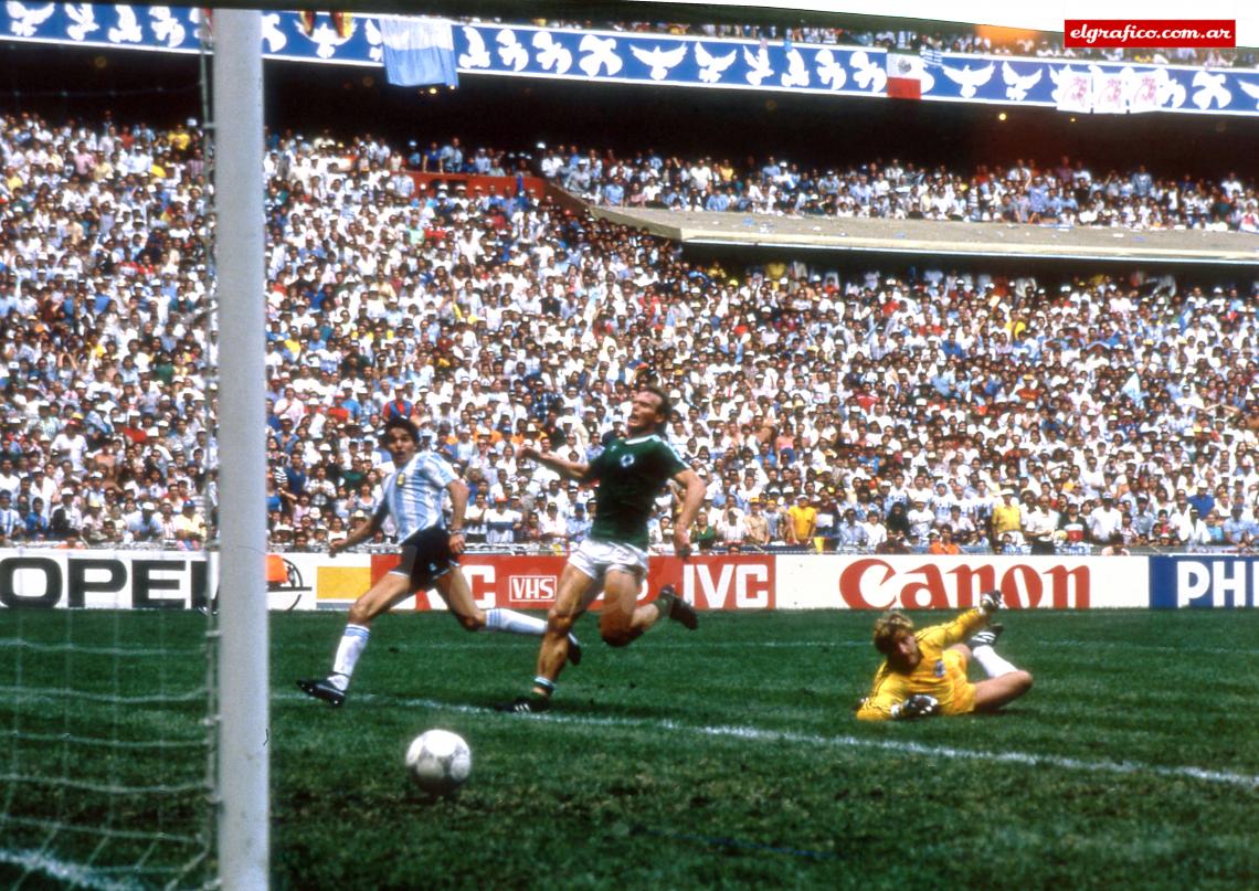An Argentine soccer player scores a goal past a diving West German goalkeeper during a crowded stadium match, with fans filling the stands and advertising boards visible around the field.