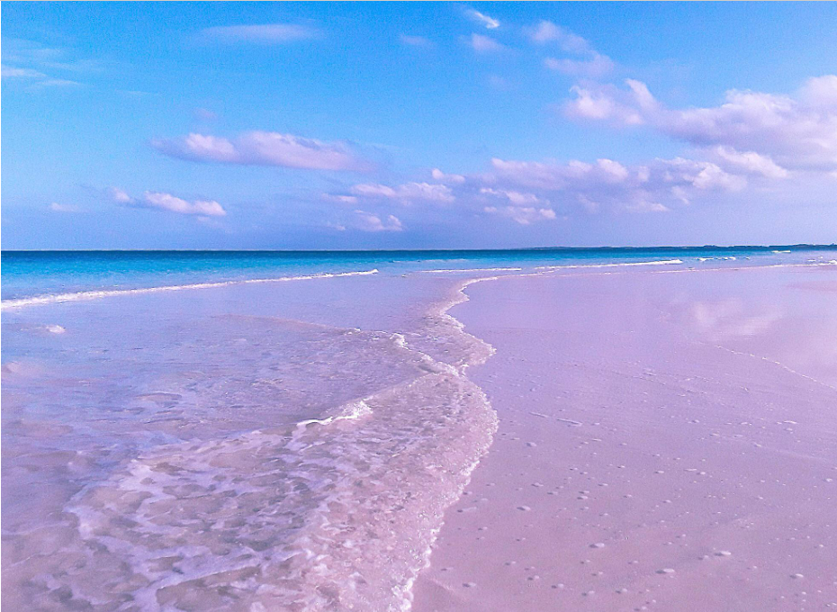 Pink-tinted sand beach with gentle waves meeting the shore under a blue sky, scattered with soft clouds. The calm ocean stretches to the horizon, creating a serene, tranquil coastal scene.