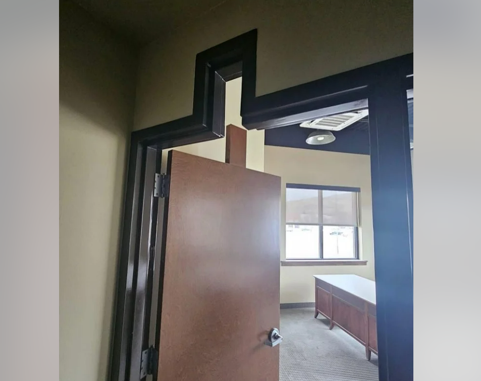 An interior office door is partially open, revealing that the door's top edge and the doorway have an unusual zigzag shape to fit around a ceiling beam. Sunlight is coming through the windows of the office beyond.