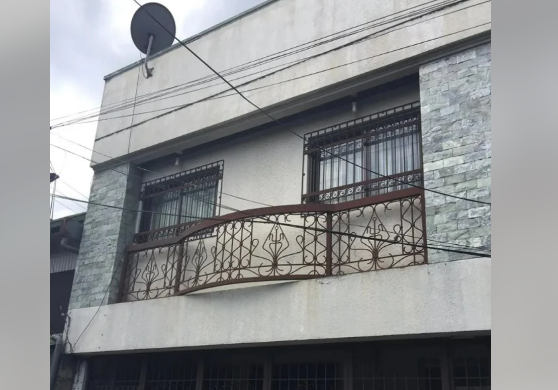 A two-story building with a balcony featuring decorative metal railing, barred windows, stone accent walls, and a satellite dish mounted near the roof. Power lines cross in front of the building.