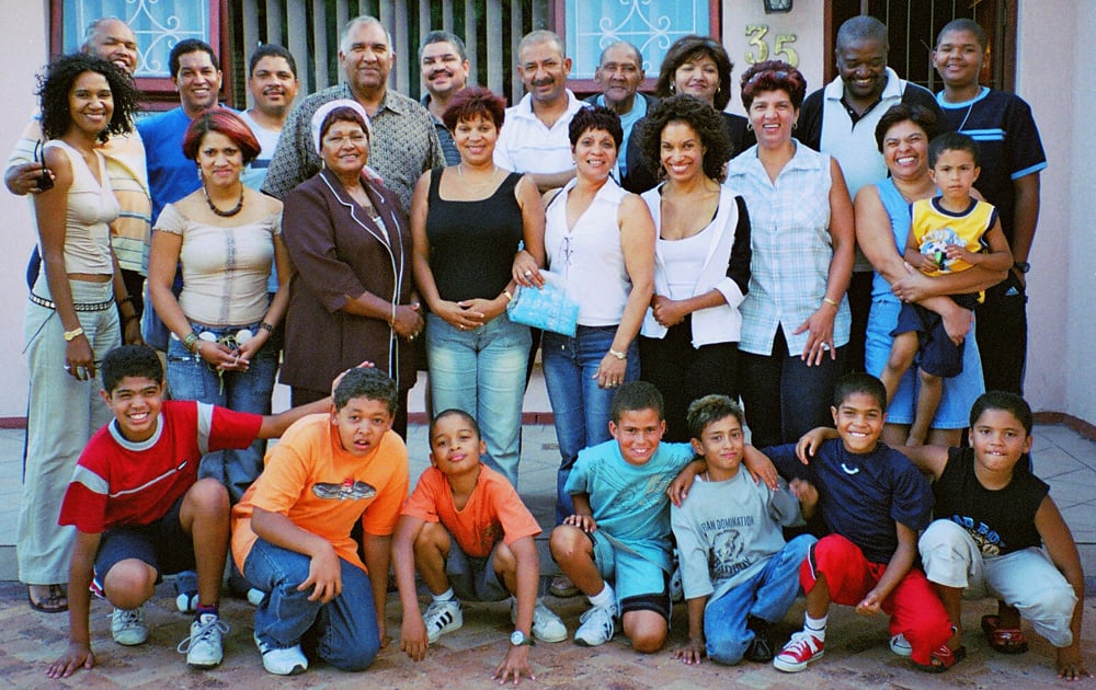 A large group of people, including men, women, and children, pose together and smile outside a building. Some adults stand at the back while children and a few adults kneel or sit in the front row.