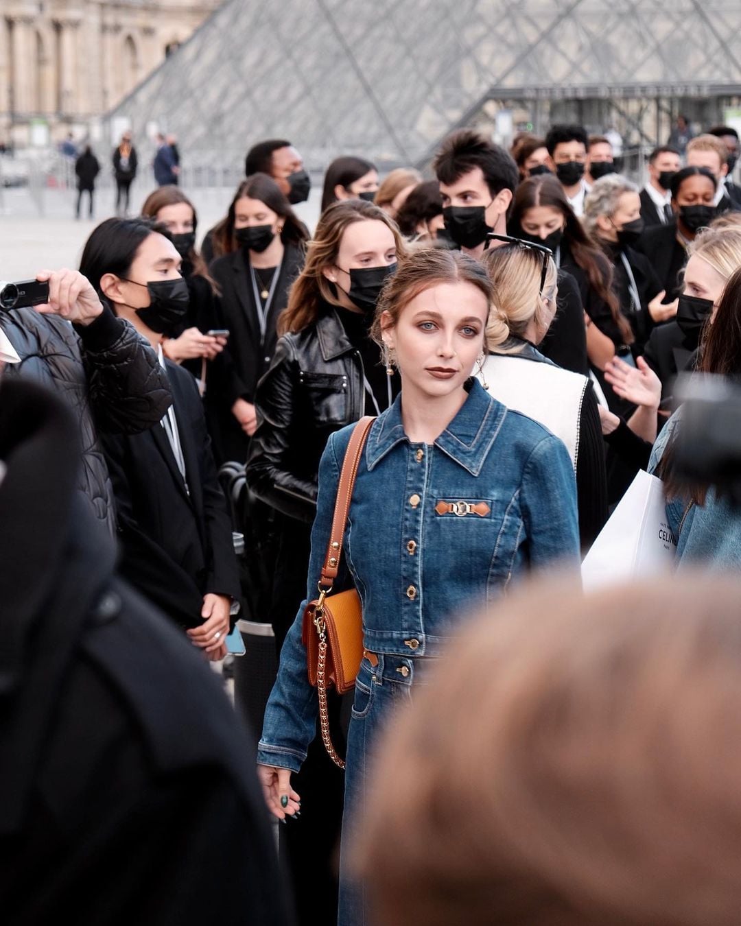 A woman in a denim dress with a brown shoulder bag stands in a crowd of people, many wearing black clothing and face masks, outside the glass pyramid entrance of the Louvre Museum in Paris.
