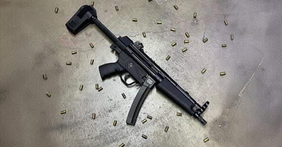 A black submachine gun with a curved magazine lies on a concrete surface, surrounded by scattered brass-colored bullets.