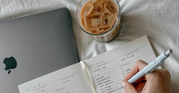 An open notebook with handwritten lists, a silver laptop, and a glass of iced coffee are on a white surface. A hand is writing in the notebook with a pen.