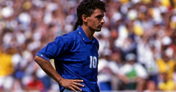 A male soccer player in a blue jersey with the number 10 stands on the field with his hands on his hips, looking focused. A blurred crowd fills the background.