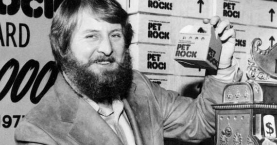 A man with a beard and mustache holds up a small Pet Rock box and smiles. Stacks of boxes labeled "Pet Rock" are visible in the background. The image is black and white.