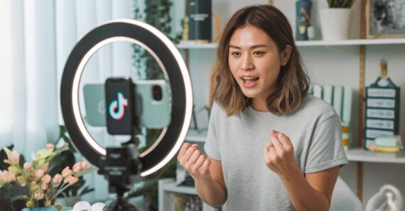 A woman sits in front of a ring light and smartphone with the TikTok logo, speaking enthusiastically and gesturing with her hands while recording a video in a well-lit room with plants and shelves.