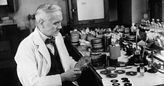 A scientist in a lab coat sits at a cluttered laboratory bench, holding a petri dish and examining its contents. Numerous petri dishes, bottles, and lab equipment are spread out on the table around him.