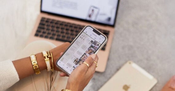 A person wearing gold bracelets and rings holds a smartphone displaying a social media feed. An open laptop and a tablet, both gold-colored, are on a light gray surface in the background.