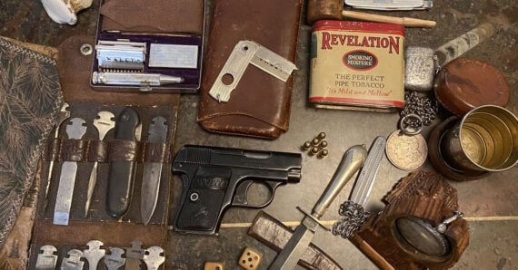 A collection of vintage items on a table, including a small handgun, pocket knives, pipe tobacco tin, coins, bullets, leather pouches, tools, dice, a folding ruler, and a pocket watch.