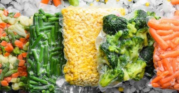 Assorted frozen vegetables in clear bags, including mixed vegetables, green beans, corn, broccoli, and baby carrots, lying on a bed of ice.