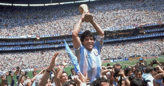 A soccer player in an Argentina jersey lifts the FIFA World Cup trophy above his head while surrounded by cheering teammates, photographers, and a stadium full of spectators.