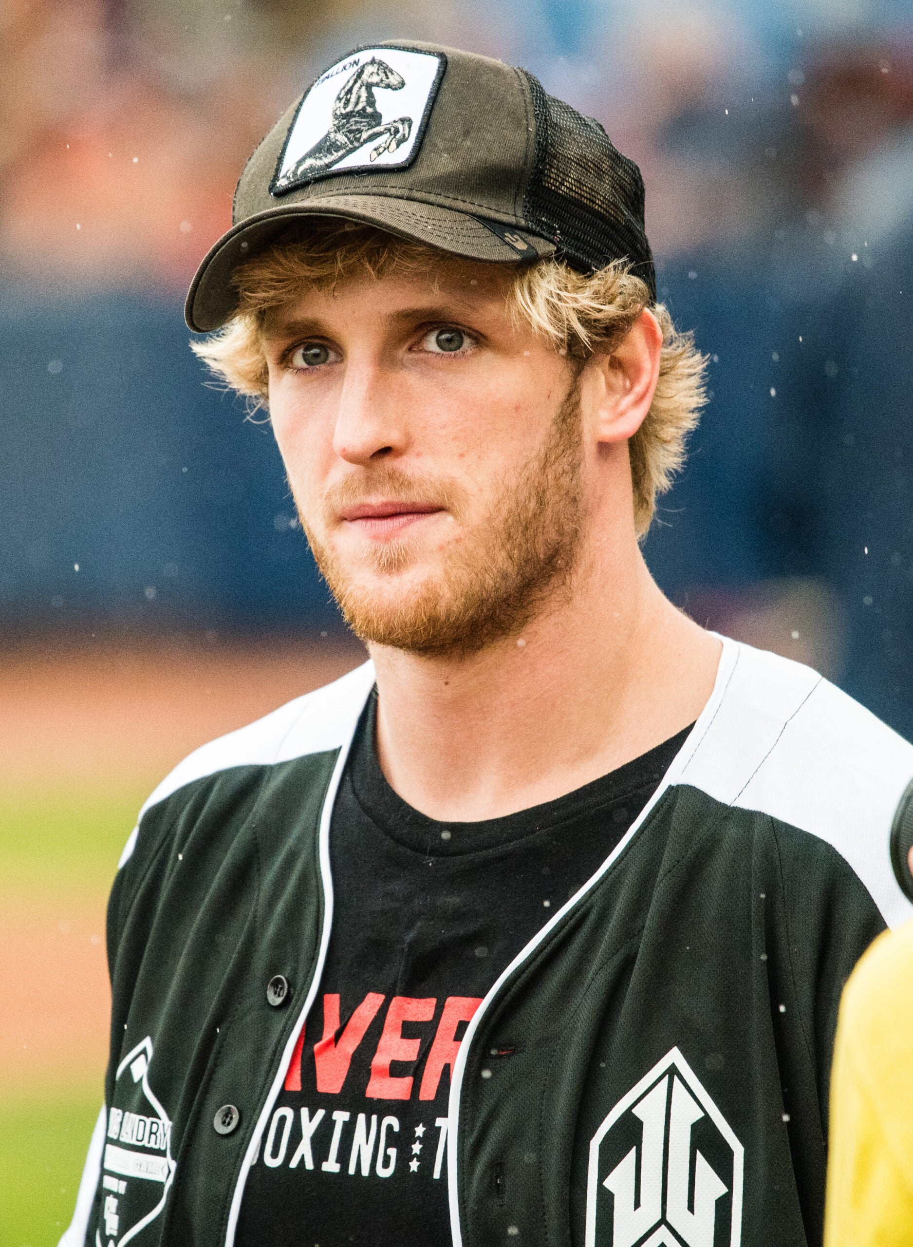 A young man with blonde hair and a beard wears a black and white baseball jersey, a black T-shirt, and a black trucker hat with a white patch, standing outdoors on what appears to be a sports field.
