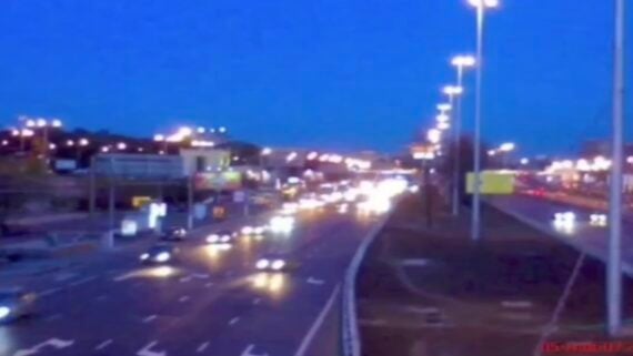 A blurry photo of a busy multi-lane highway at dusk, with numerous cars and bright streetlights illuminating the scene. Buildings and signs line the roadside under a deep blue sky.