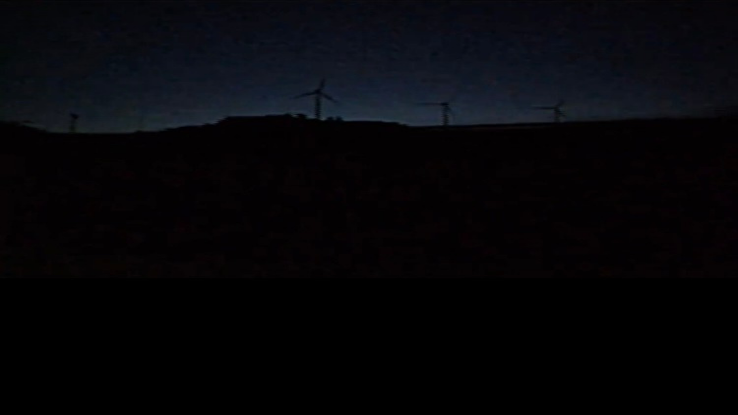 A dark landscape at night shows wind turbines silhouetted against a faintly glowing sky, with a hill barely visible in the darkness.