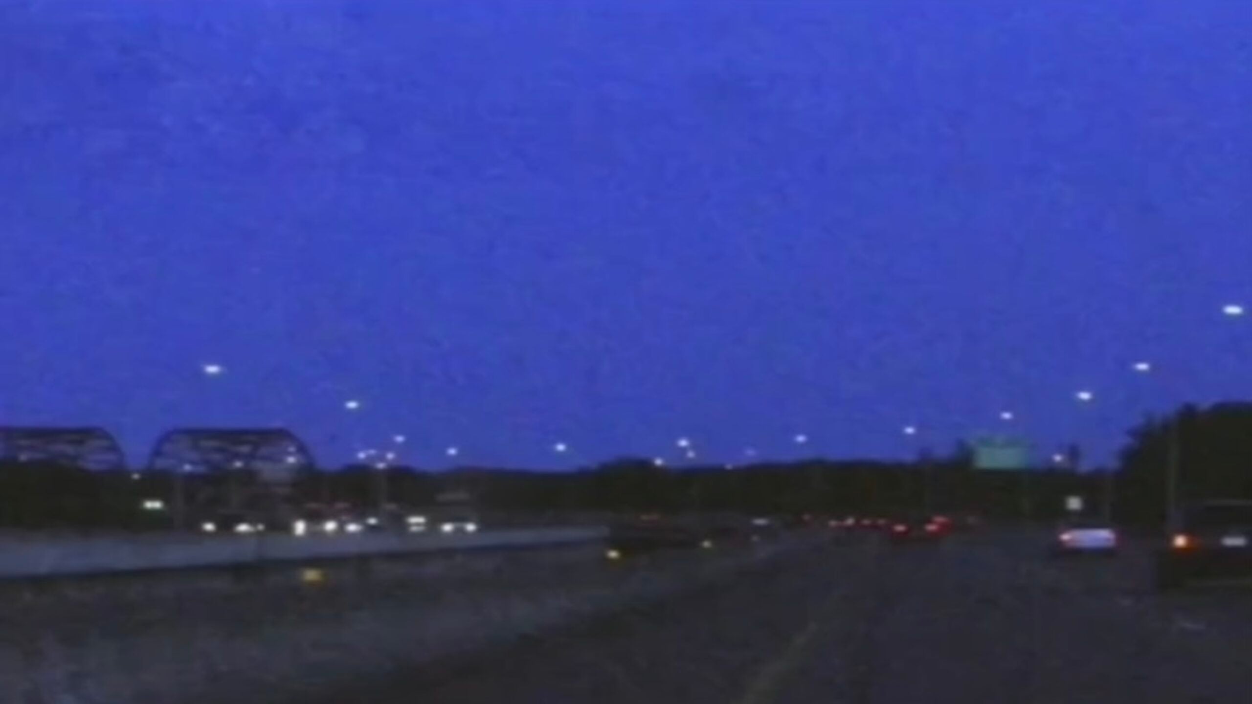 A nighttime scene of a highway shows cars driving with headlights on, streetlights illuminating the road, and a bridge structure visible on the left under a dark blue sky.