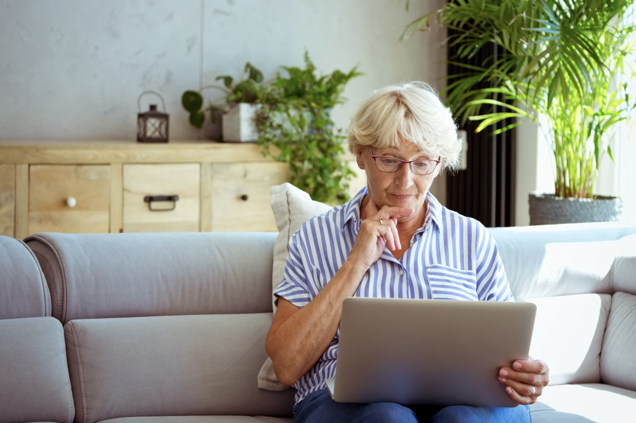 An older woman with short gray hair and glasses sits on a gray sofa, looking thoughtfully at a laptop. She is wearing a striped shirt, and there are green plants and wooden furniture in the background.