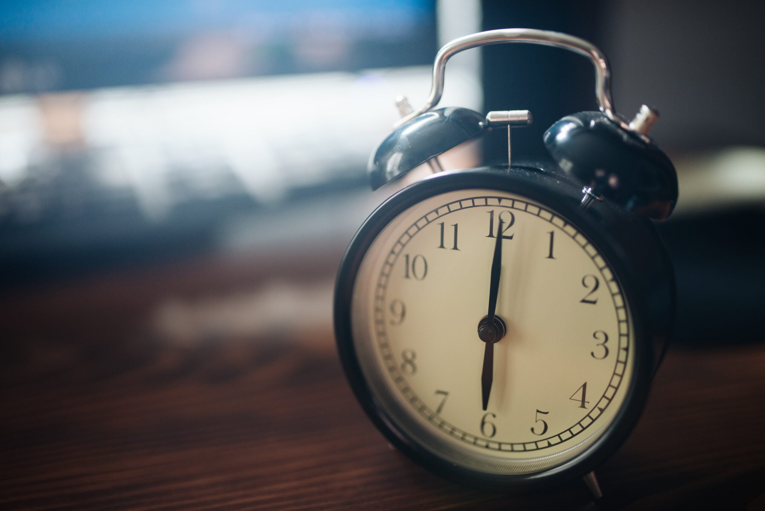 A close-up of a black analog alarm clock showing 7:00, placed on a wooden surface with a blurred background, possibly a computer or laptop.