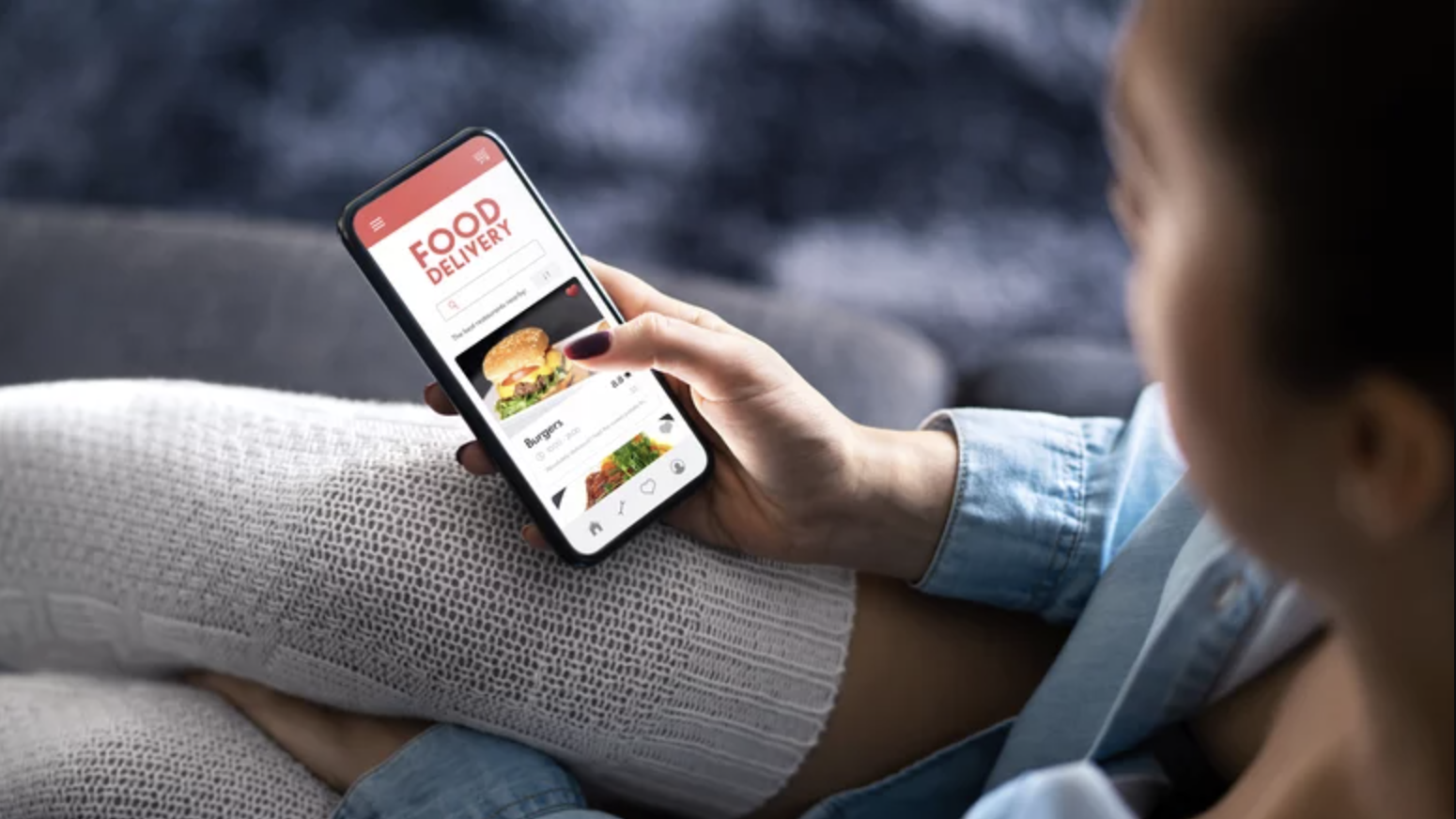 A person sitting on a couch, wearing a blue shirt and white knit socks, using a smartphone to browse a food delivery app that displays images of burgers and other meal options.