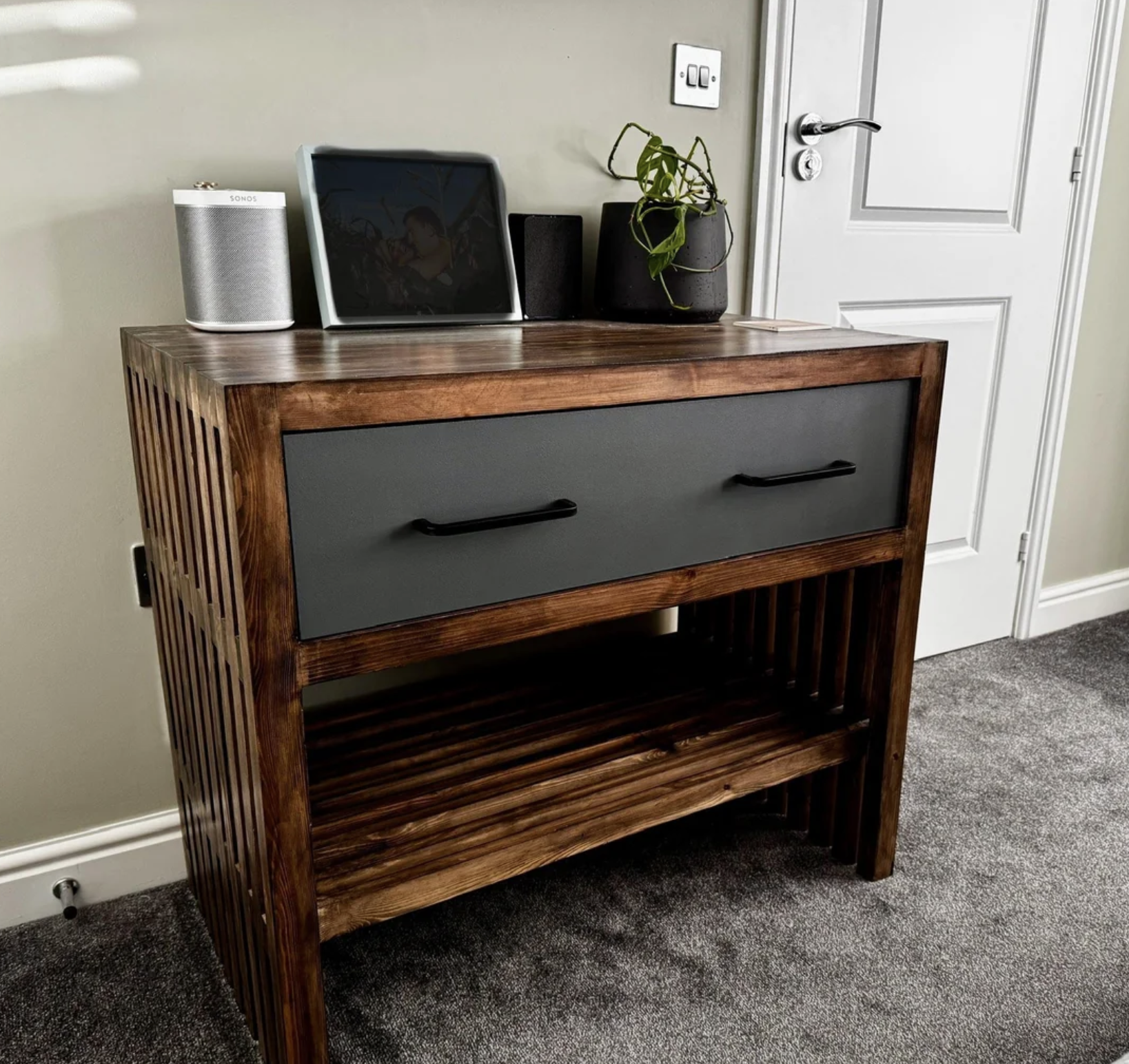 A wooden side table with slatted sides and a single drawer holds a smart speaker, a tablet with a dark screen, and a small potted plant. The table stands on a gray carpet near a closed white door.