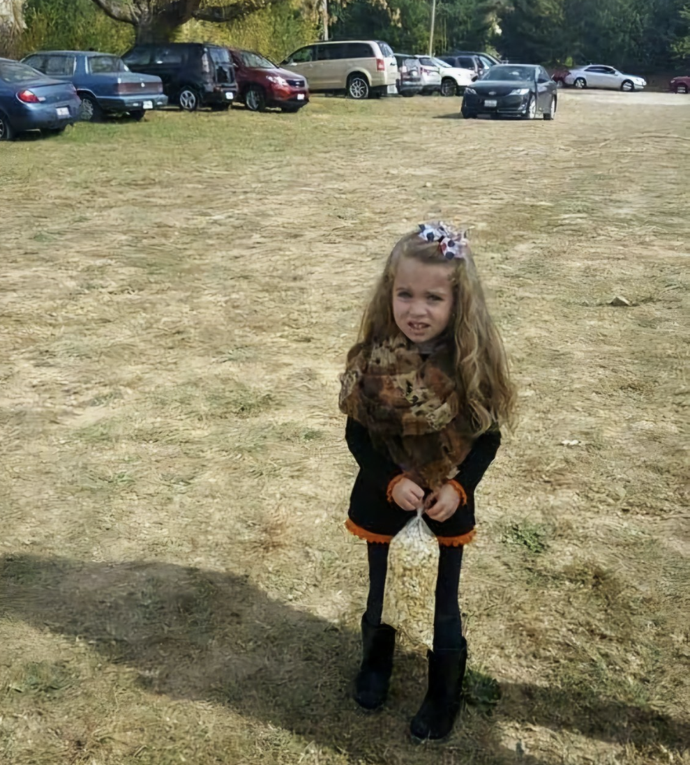 A young girl with long curly hair, wearing a scarf, dark dress, and boots, stands on dry grass holding a bag of popcorn. Several parked cars and trees are visible in the background.