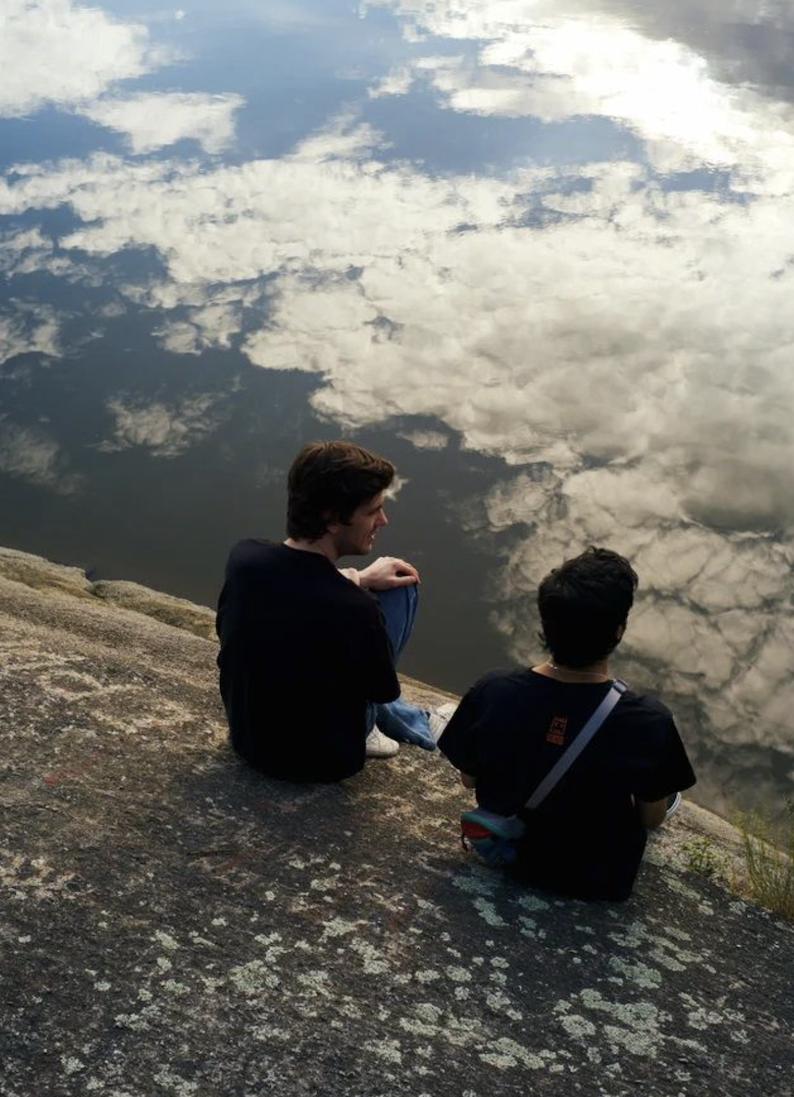 Two people sit on a rocky surface by calm water that reflects the sky and clouds, creating an illusion as if they are sitting among the clouds.