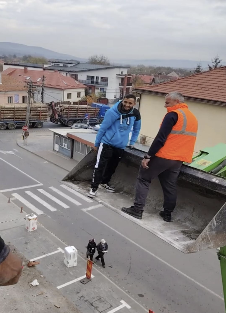 Two men stand in the bucket of a raised construction vehicle above a street, one wearing a blue hoodie and the other an orange safety vest. Buildings, parked trucks, and people walking are visible below.