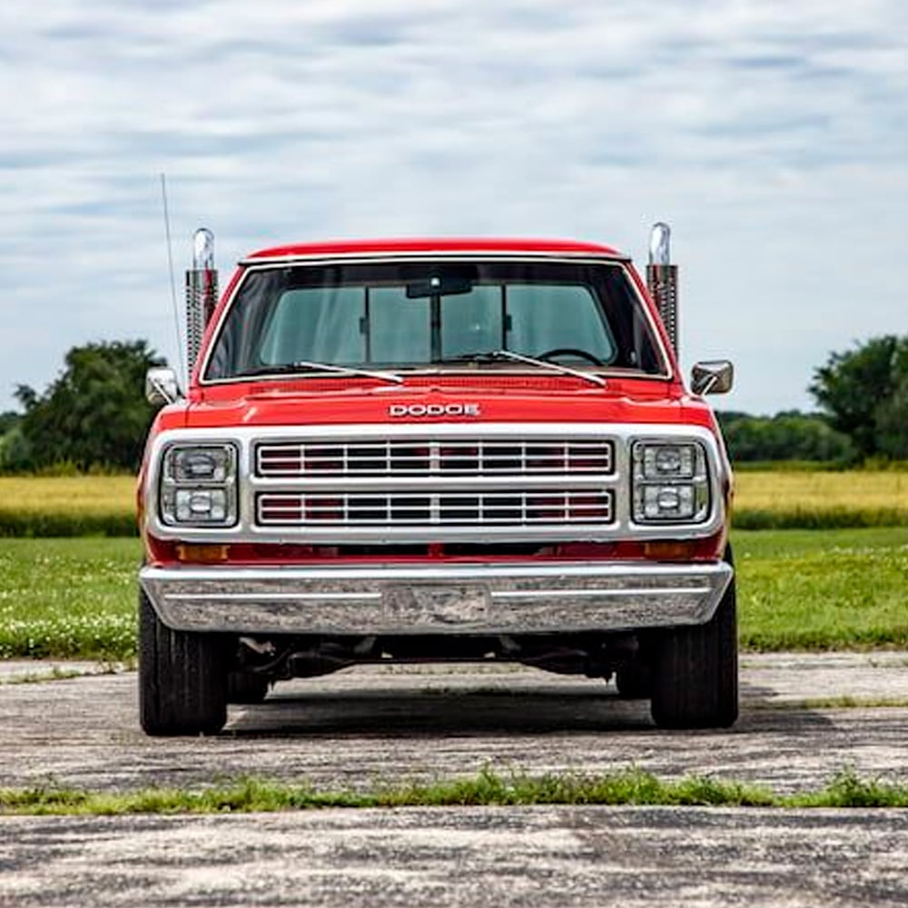 A red vintage Dodge pickup truck is parked on a paved surface, facing the camera, with green grass and trees in the background under a partly cloudy sky.