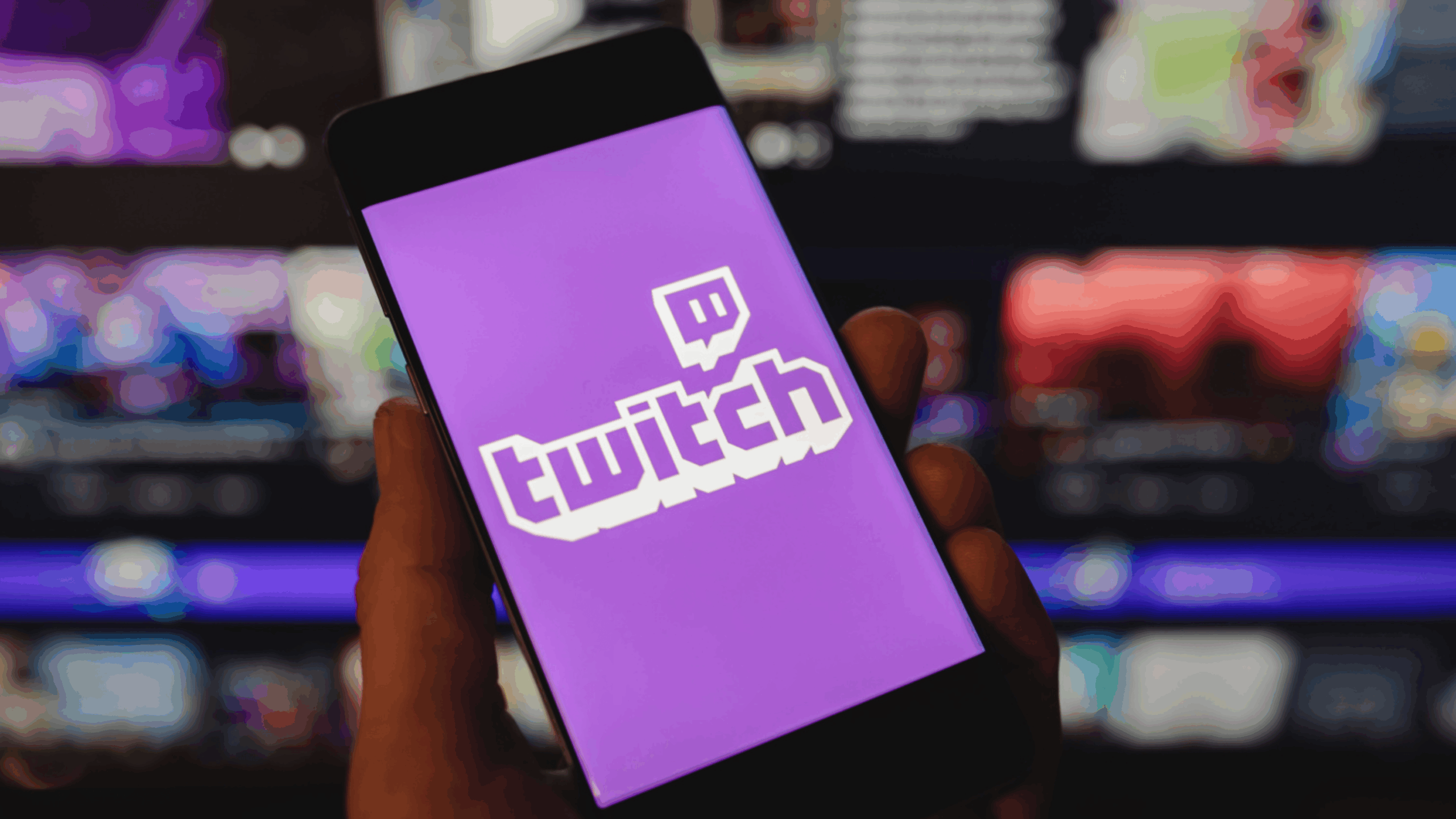 A hand holds a smartphone displaying the Twitch logo on a purple background, with a blurred computer screen showing various content in the background.