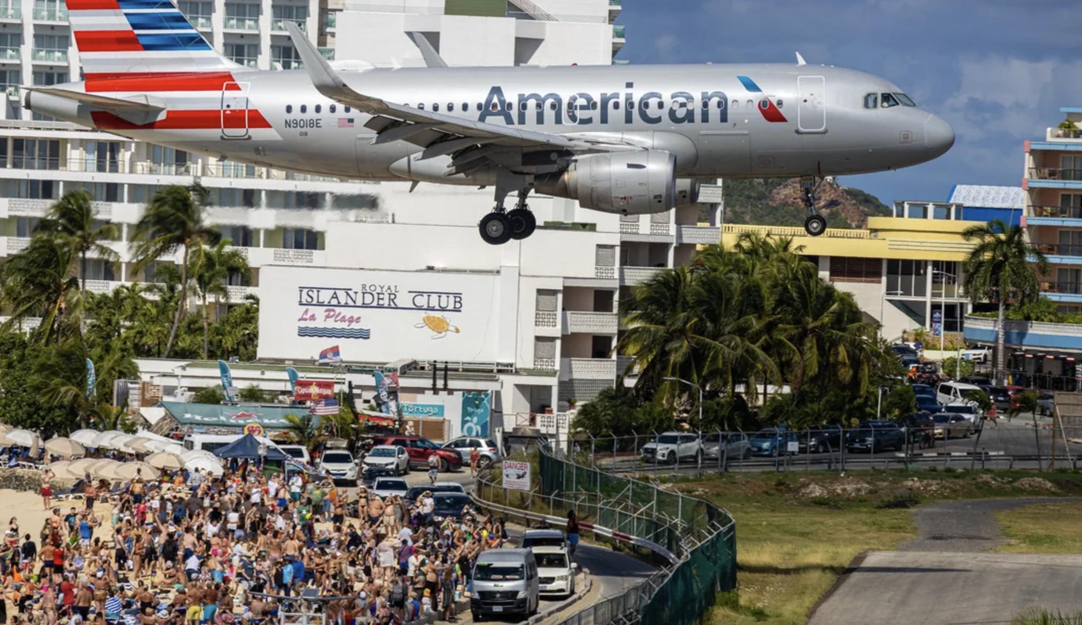 A large American Airlines plane flies very low over a crowded beach and buildings, preparing to land near Maho Beach in St. Maarten, as people on the sand watch and take photos.