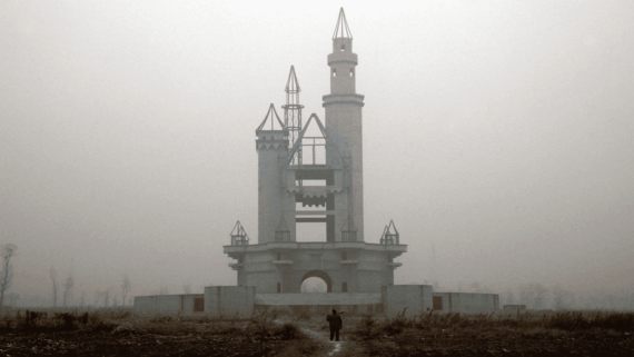 A person stands in front of an unfinished, abandoned castle structure surrounded by barren land and fog, giving the scene an eerie, desolate atmosphere.