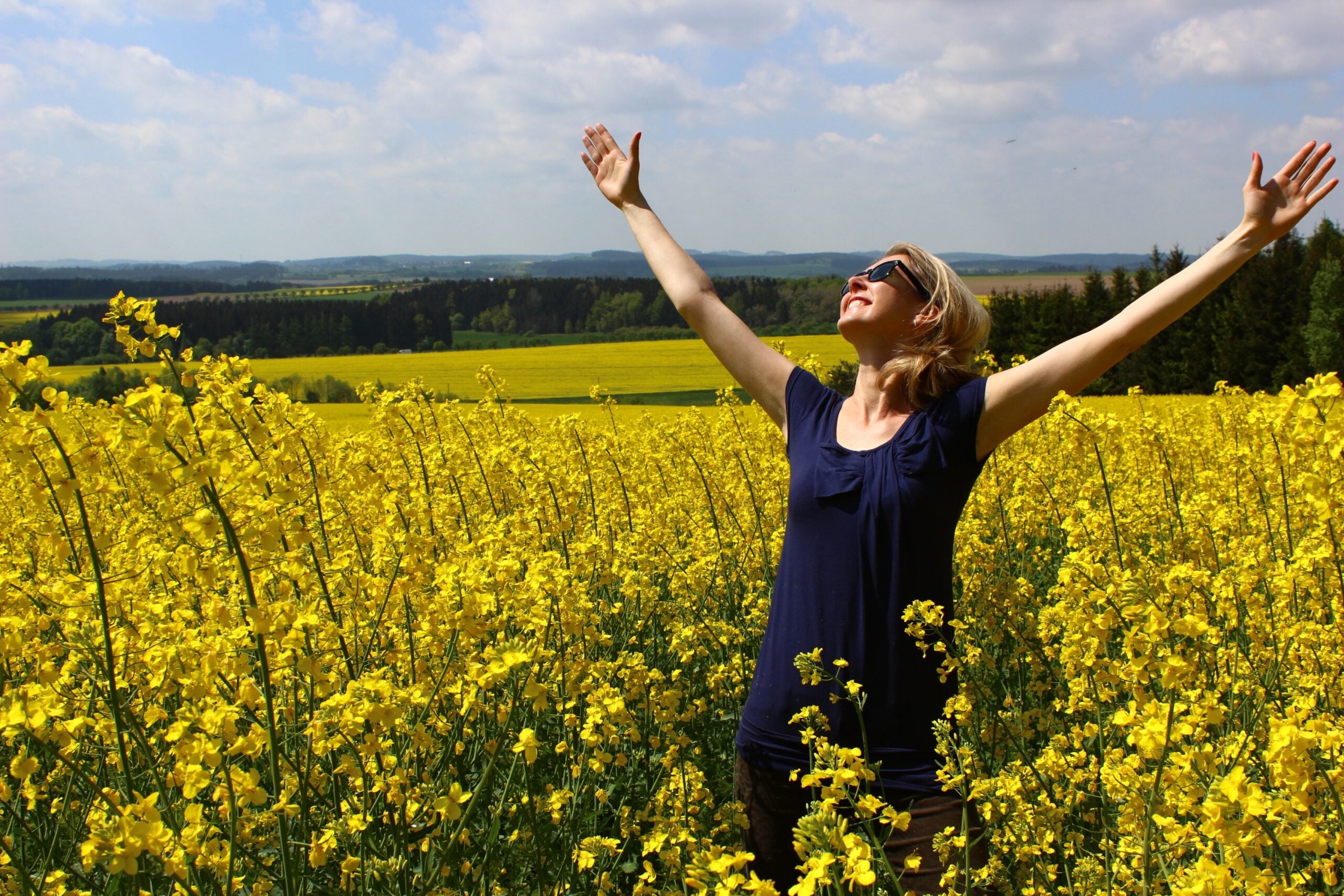 A woman stands in a field of yellow flowers with her arms raised and eyes closed, appearing joyful and relaxed under a partly cloudy sky. Rolling hills and trees are visible in the background.