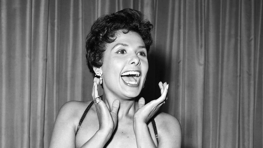 A woman in a sleeveless dress smiles widely with her mouth open and hands raised near her face, standing in front of a curtain.