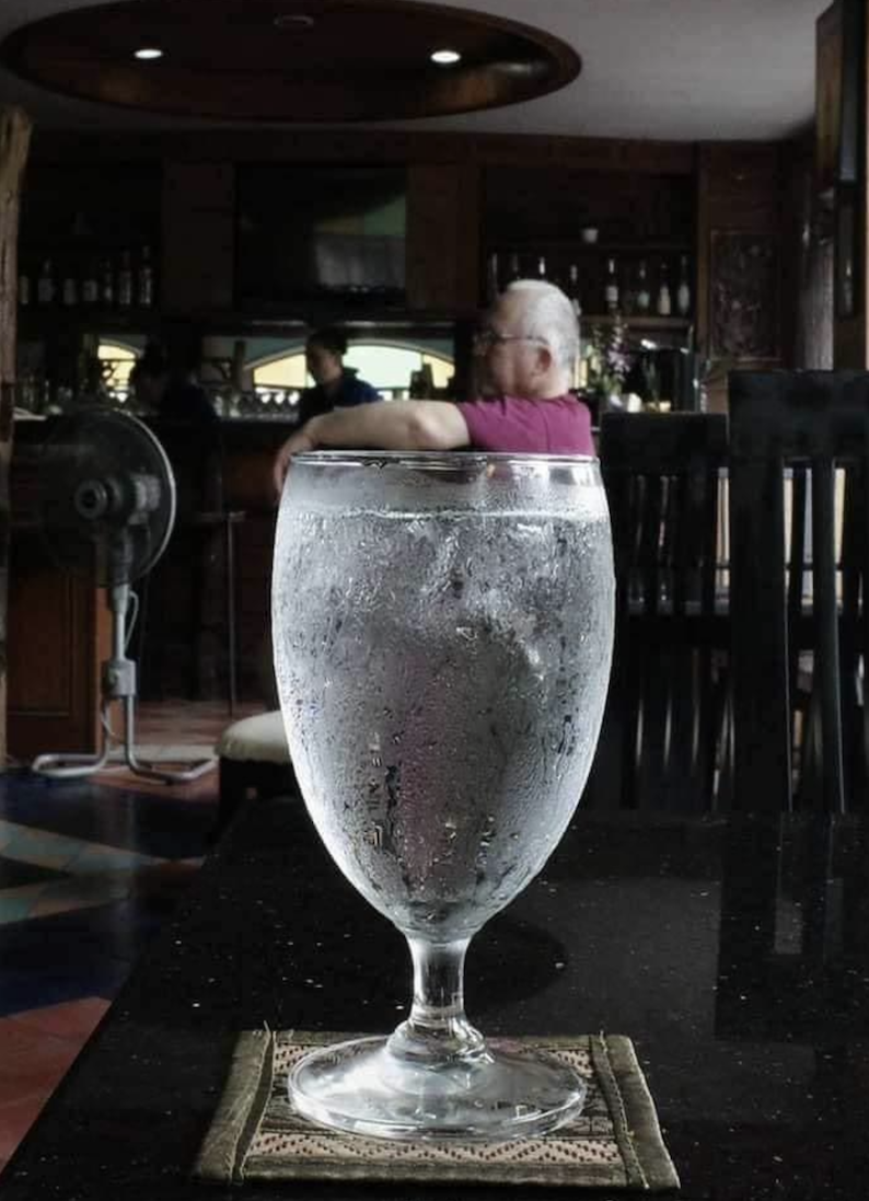A close-up of a cold, frosted glass of water on a coaster, placed on a dark table in a restaurant, with a man in a purple shirt sitting in the blurred background.