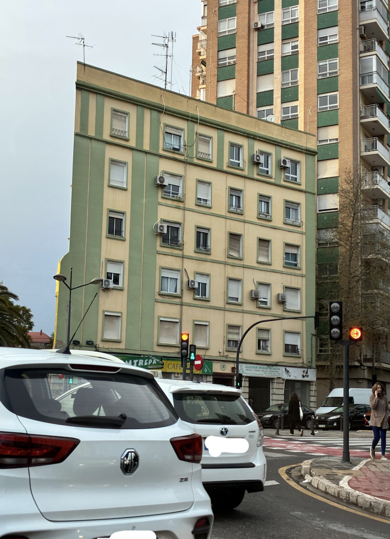 A city street with cars waiting at a red traffic light in front of a green and yellow apartment building. Pedestrians are crossing and taller buildings are visible in the background.