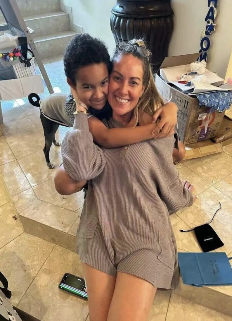 A young child hugs a smiling woman in a beige knit outfit indoors. A dog stands nearby, and open boxes are visible in the background, suggesting a celebration or move.