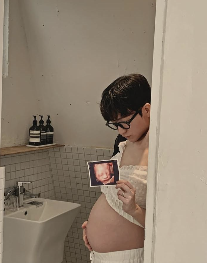 A pregnant person wearing glasses and a white outfit stands in a bathroom, holding an ultrasound photo of a baby near their bare belly.