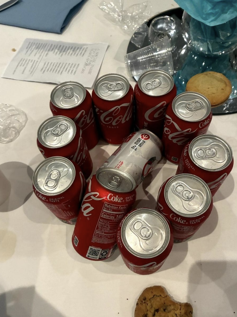 Thirteen red Coca-Cola cans form a circle around a single Diet Coke can on a table with cookies, napkins, and a blue decorative item in the background.