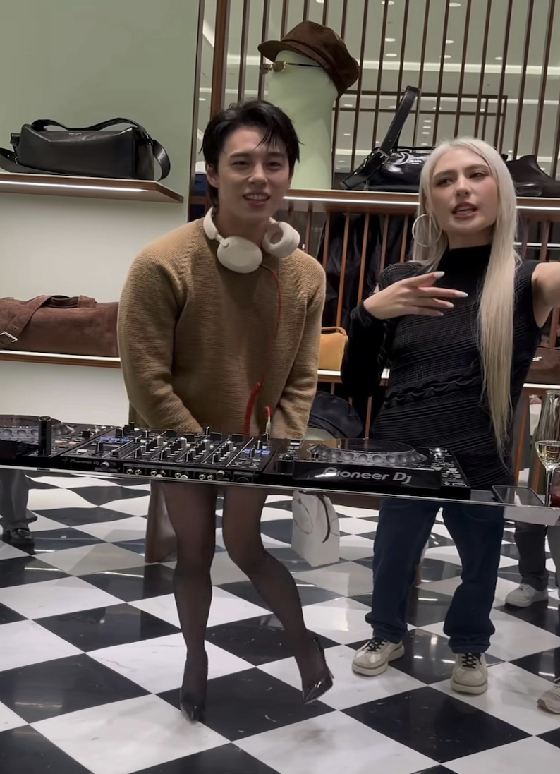Two people stand in a stylish store with checkered floors and shelves of bags. One, wearing a sweater, headphones, and sheer tights, stands behind DJ equipment; the other, with long blond hair, gestures with one hand.