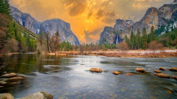 A river flows through a valley surrounded by tall mountains and pine trees at sunset, with a dramatic orange sky and clouds illuminating the landscape.