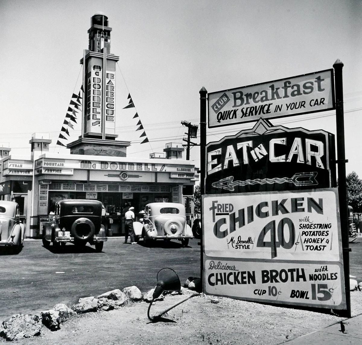 Black and white photo of a vintage drive-in restaurant with cars parked in front; a large sign advertises fried chicken for 40¢ and chicken broth for 10¢ or 15¢, with the slogan "EAT IN CAR.