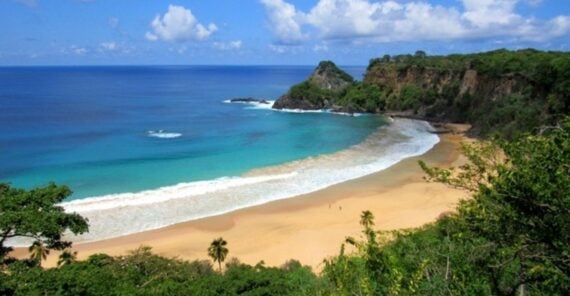 A scenic view of a tropical beach with golden sand, turquoise water, gentle waves, lush green cliffs, and a partly cloudy blue sky in the background.
