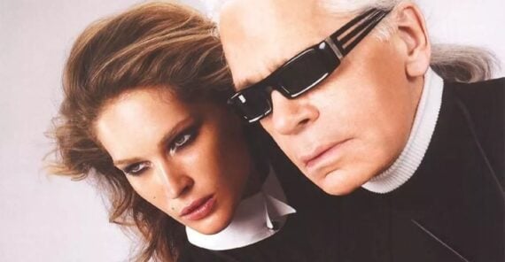 A woman with voluminous brown hair and dramatic makeup poses closely beside an older man with long white hair, wearing black sunglasses and a black turtleneck. Both appear serious and stylish against a pale background.