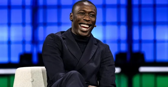 A man in a dark suit sits on a chair, smiling broadly during an event. The background features a blue and green geometric pattern.