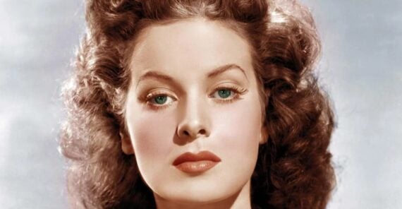 A woman with wavy brown hair, fair skin, blue-green eyes, and red lipstick looks directly at the camera against a light background.