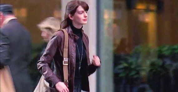 A woman with long brown hair wearing a brown jacket and carrying a beige shoulder bag walks confidently outdoors in an urban setting, with blurred people and buildings in the background.