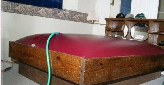 A waterbed with a maroon mattress is overfilled and swollen, stretching above the wooden frame. A green hose is attached to the mattress, indicating it is still being filled with water. The bed is in a carpeted room with shelves and decor.