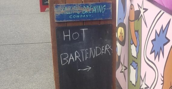 A chalkboard sign outside a brewery reads &ldquo;HOT BARTENDERS &rarr;&rdquo; in white chalk, with a colorful mural and sidewalk visible in the background.